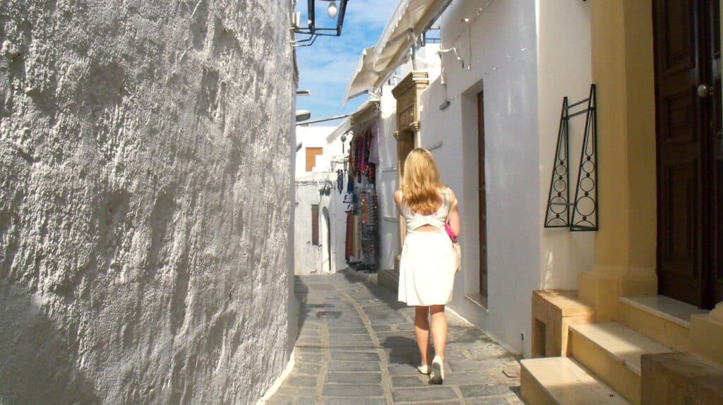 A solo female traveller in a white dress walks down a narrow, sunny street with white-washed walls and stone pavement.