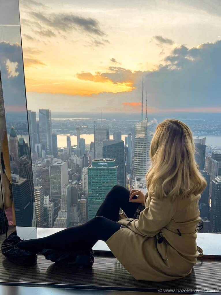 Woman with blonde hair sits at a floor-to-ceiling window looking out over New York City at sunset.