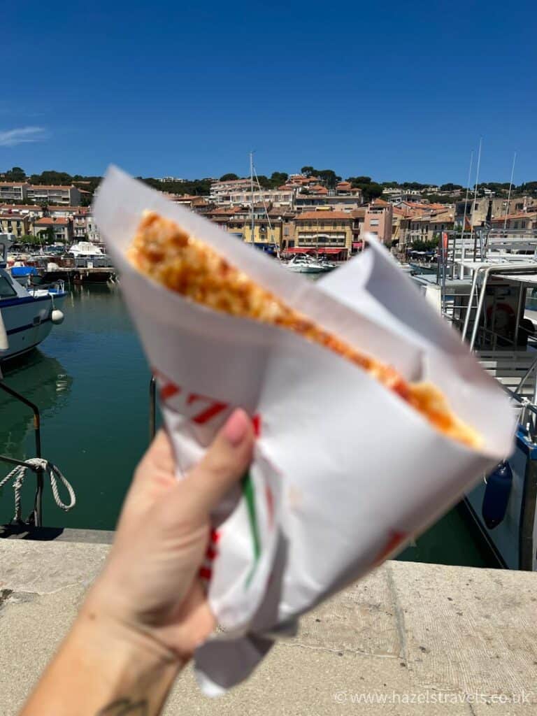 Hand holding a folded slice of takeaway pizza in front of the Cassis harbour, with colourful buildings and boats in the background on a sunny day.