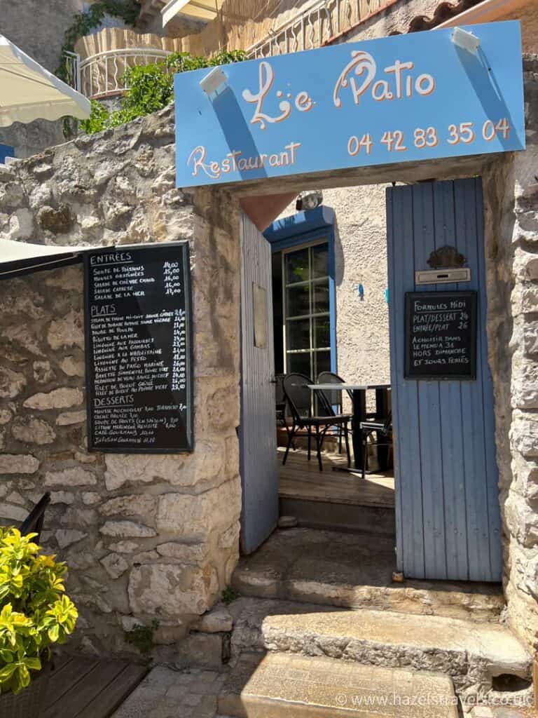Stone entrance to a rustic French restaurant in Cassis, with a blue wooden door, chalkboard menus, and a blue sign reading “Les Belles Canailles.”