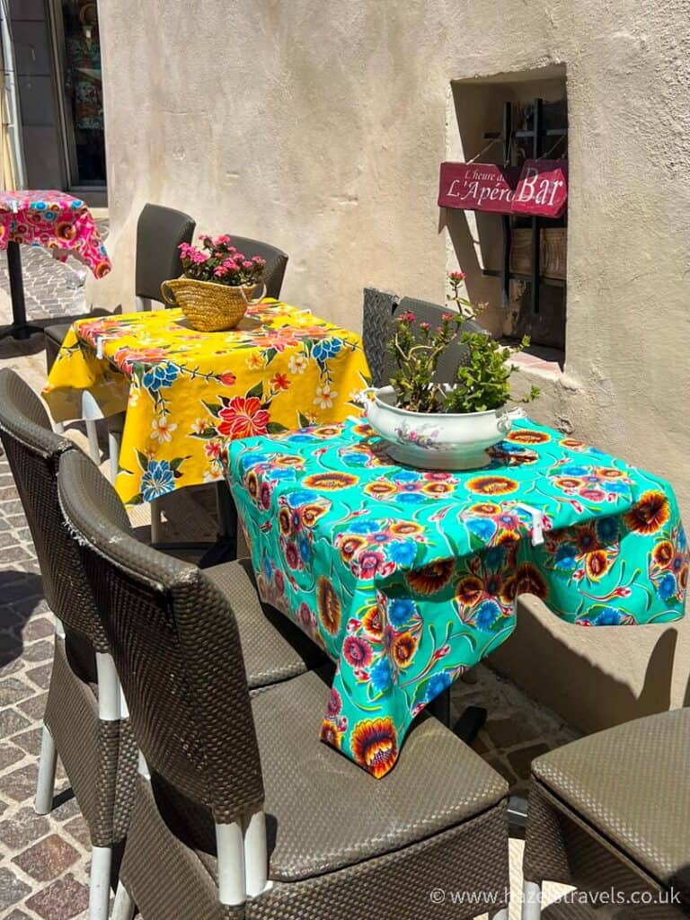 Outdoor café tables in Cassis covered with colourful Provençal tablecloths, set against a cream stucco wall with potted flowers and cobblestone flooring.