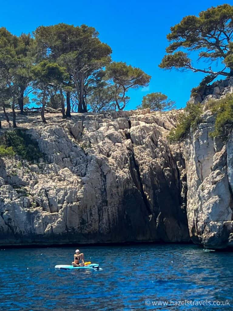 Person paddleboarding on deep blue water beneath white limestone cliffs and pine trees in the Calanques near Cassis, France.