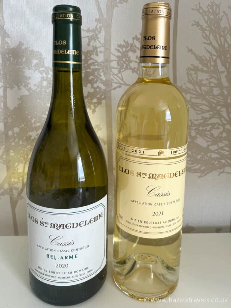 Two bottles of white wine from Cassis, France, displayed against a soft neutral background with delicate botanical wallpaper.