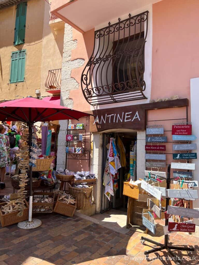 Charming souvenir shop in Cassis, France, with postcards, baskets, and textiles displayed outside under a red parasol on a sunny cobbled street.