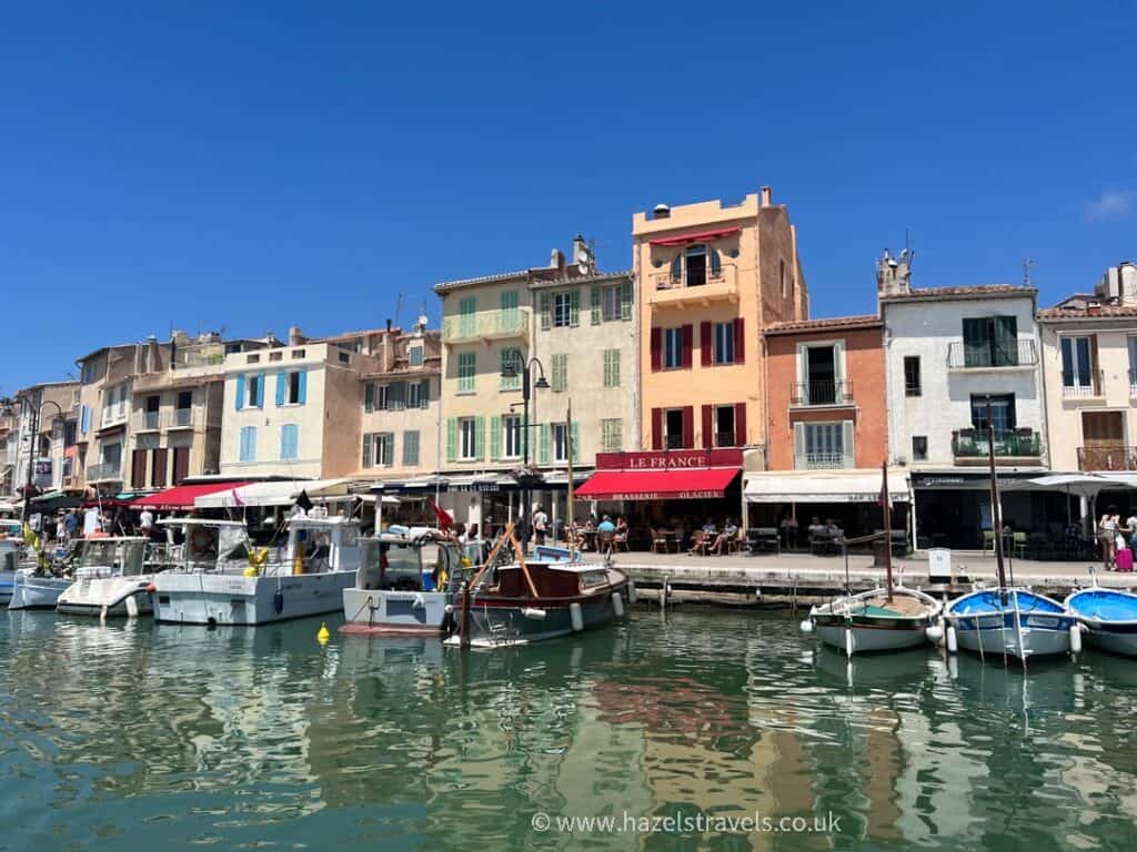 Colourful harbourfront buildings in Cassis, France, reflected in the calm water with small boats moored along the quay on a bright sunny day.