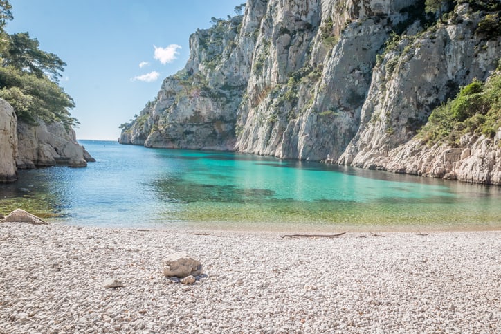 Pebble beach leading to turquoise water and steep limestone cliffs at Calanque d’En-Vau near Cassis, with sunlight reflecting off the sea.