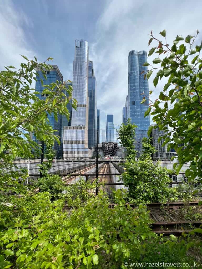 Tall modern skyscrapers of Edge NYC peek through lush green foliage and a wire fence, as railway tracks lead the way towards the vibrant city.