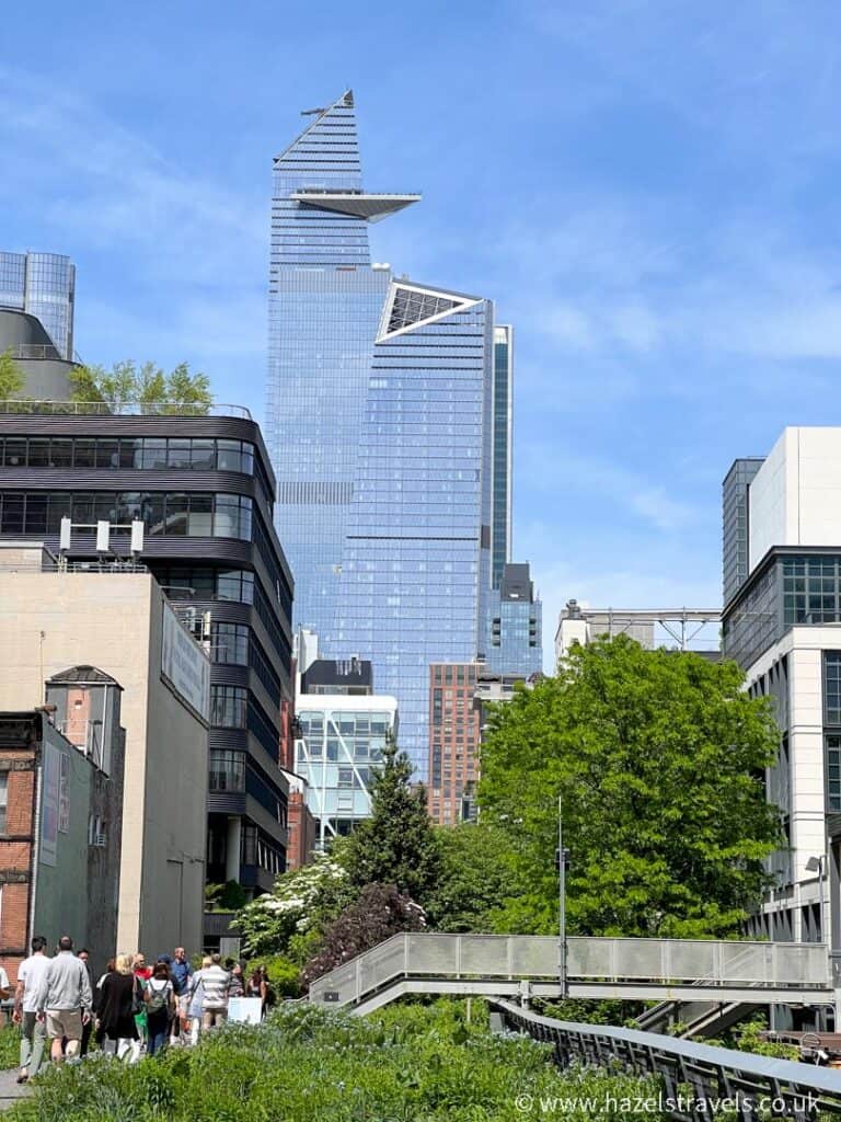 The angular design of the tall glass skyscraper stands prominently above the shorter buildings and greenery, reminiscent of iconic structures like Edge NYC. In the foreground, people stroll leisurely along a pathway, adding life to the urban landscape.