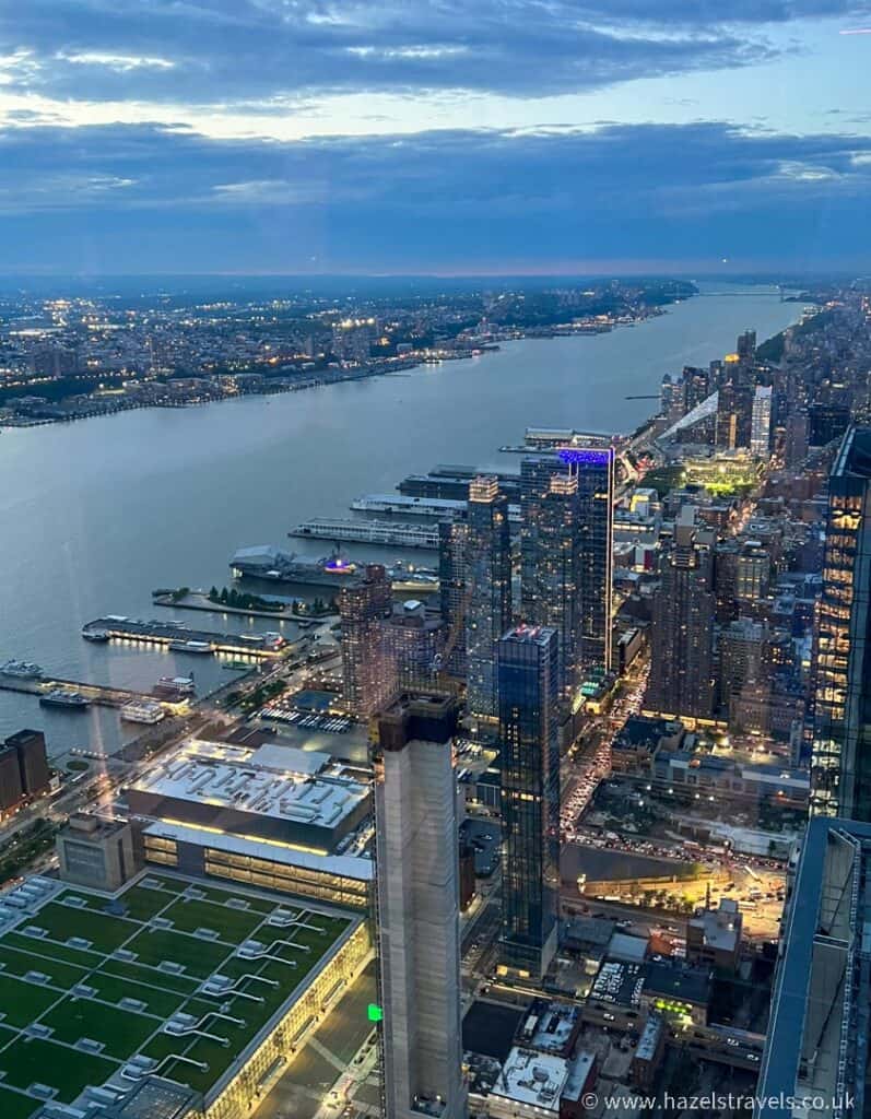 Aerial cityscape view of a bustling city at dusk with Edge NYC towering above the river on the left, tall buildings, and illuminated streets.