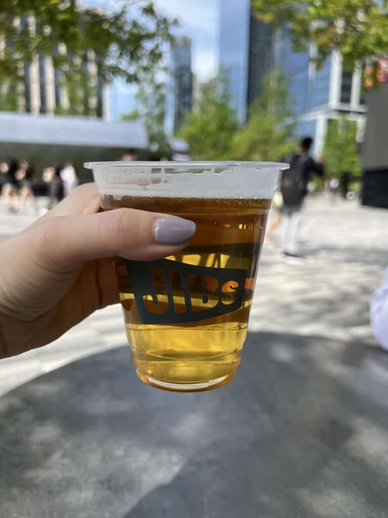 A hand holds a plastic cup of beer in an urban oasis at Hudson Yards, with trees and sleek buildings framing the background.