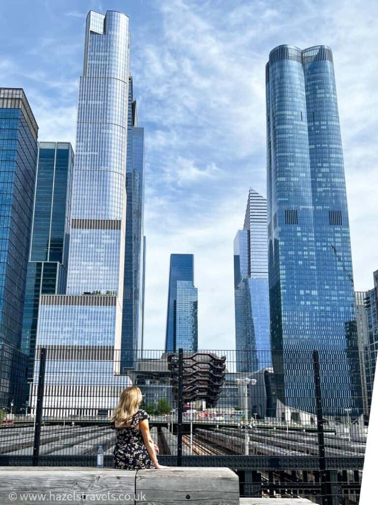 A person with long hair gazes at the Hudson Yards skyline, where tall, modern glass skyscrapers cut into a partly cloudy sky.