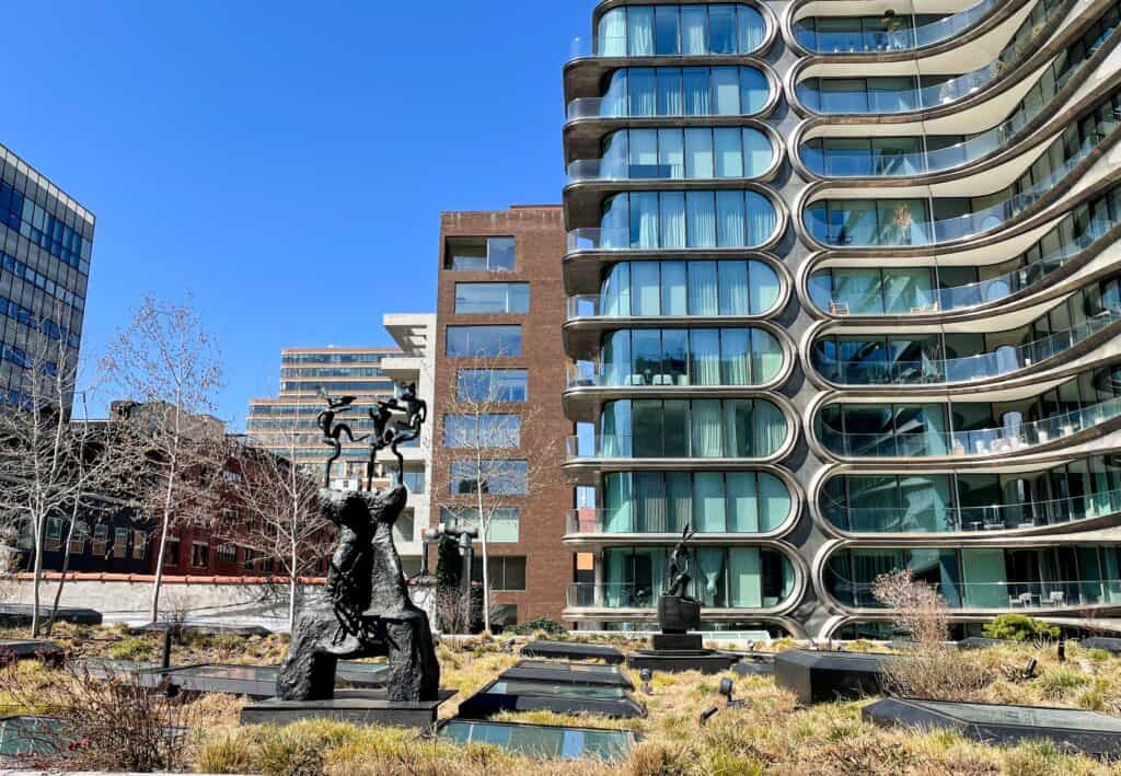 A modern building with curved glass balconies stands elegantly beside sculptures in a landscaped area, on the Highline Park NYC, under a clear blue sky.