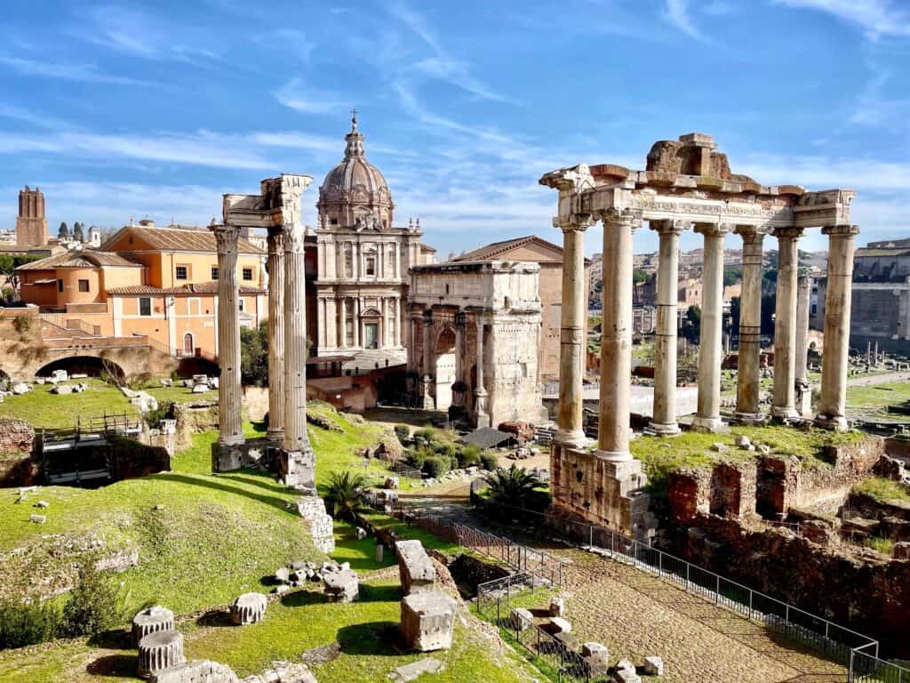 the ruins of ancient Roman columns standing amidst grassy areas and historic buildings under a clear blue sky.