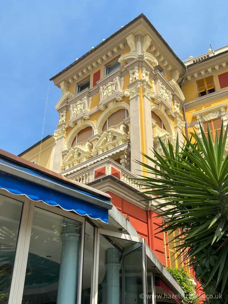 Ornate yellow and red building in Rapallo, Italy, featuring decorative stucco details, arched windows, and classical columns under a clear blue sky with palm fronds in the foreground.