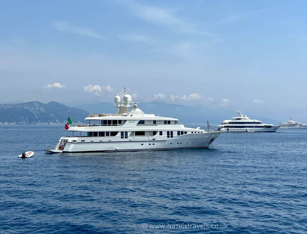 A large white yacht with an Italian flag is anchored in the calm sea near Portofino, with mountains visible in the background, offering a serene backdrop of Portofino.