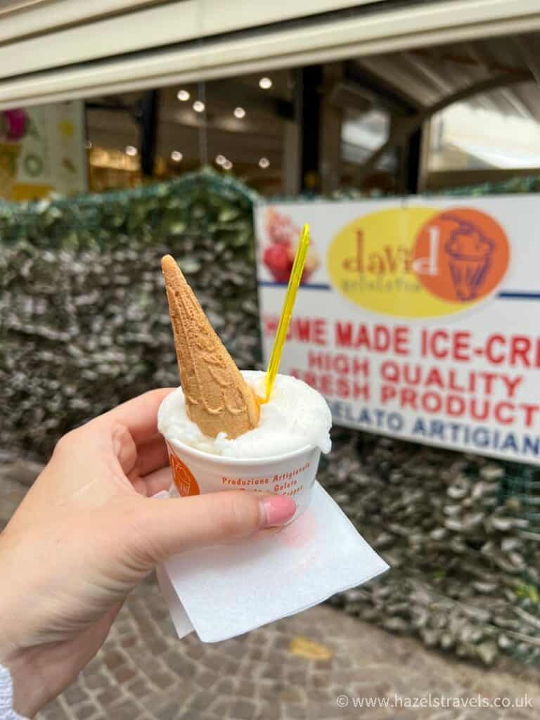 Hand holding a cup of gelato with a cone and spoon in front of an ice cream shop, one of the delightful things to do in Sorrento, Italy.