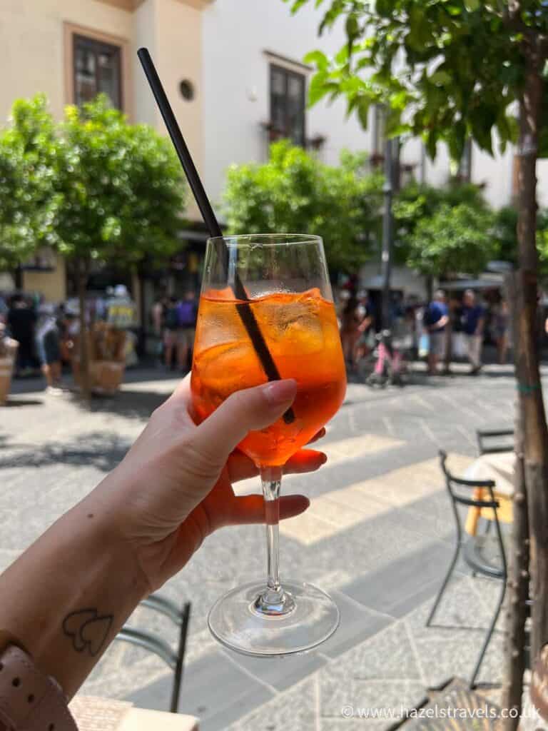 A woman holds up a glass of aperol spritz with Italian streets in the background.