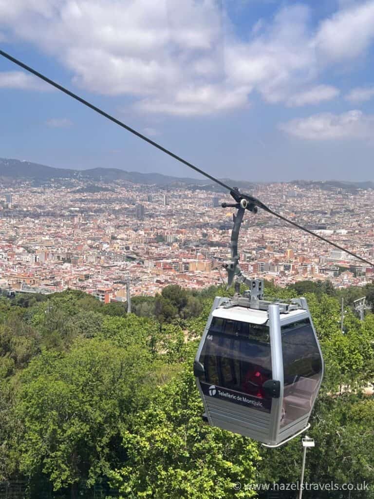 The Montjuic Cable Car gracefully glides over a lush green area, with a stunning cityscape in the background under a partly cloudy sky.