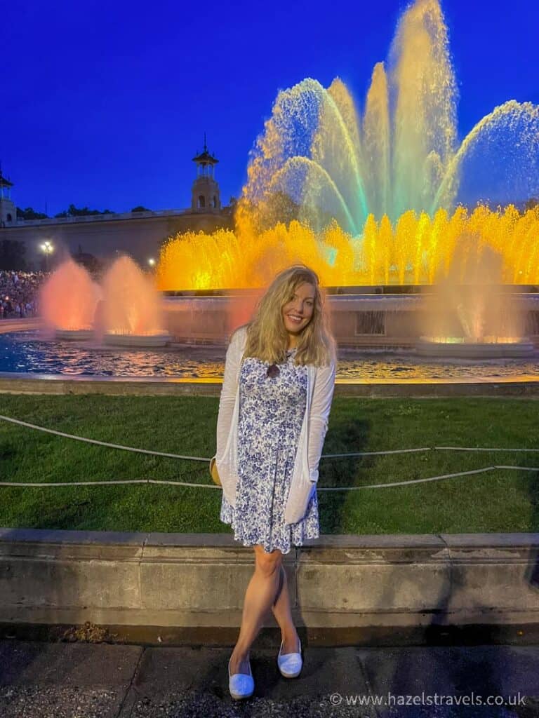 A woman stands smiling in front of the magic fountain in Barcelona, its colorful lights dancing at night. She wears a blue and white dress with a white cardigan.