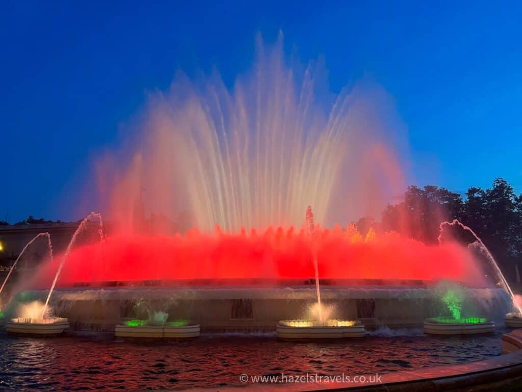 The magic fountain in Barcelona displays vibrant red and pink colors against a dark blue evening sky, creating an enchanting spectacle.