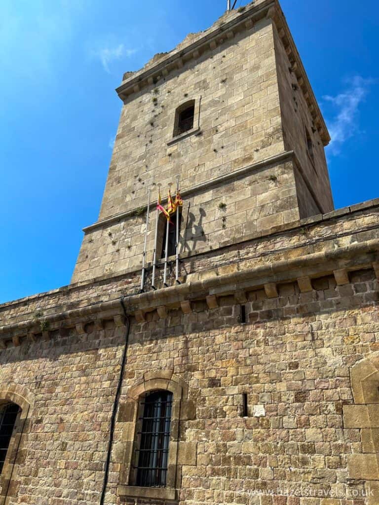 Under the clear blue sky, this stone fortress tower with barred windows stands proudly. Multiple flagpoles are attached, showcasing two vibrant flags.