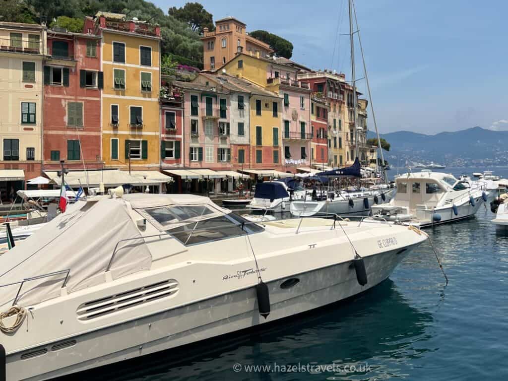 Boats moored in a scenic harbor with colorful buildings in the background under a clear sky invite you to explore the charming allure of Portofino. Among things to do in Portofino, soaking in this picturesque view is a must.