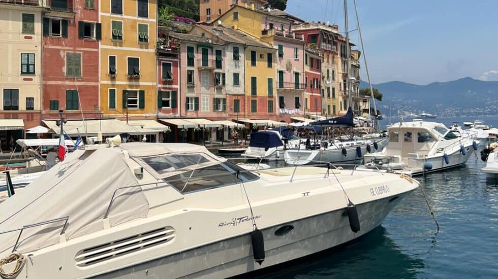 Boats moored in a scenic harbor with colorful buildings in the background under a clear sky invite you to explore the charming allure of Portofino. Among things to do in Portofino, soaking in this picturesque view is a must.