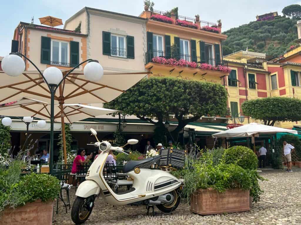 A white scooter is parked in front of vibrant cafés and colorful buildings with flower-adorned balconies, a charming snapshot of things to do in Portofino. Umbrellas and greenery enhance the picturesque cobblestone square.