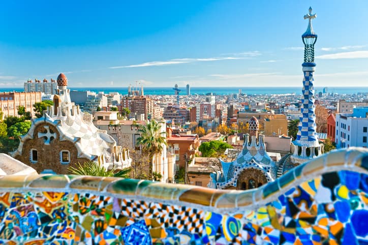 Panoramic view of Barcelona from the iconic Park Güell. The colorful mosaic structures in the foreground perfectly blend with the vibrant cityscape and shimmering sea visible in the background under a clear blue sky.