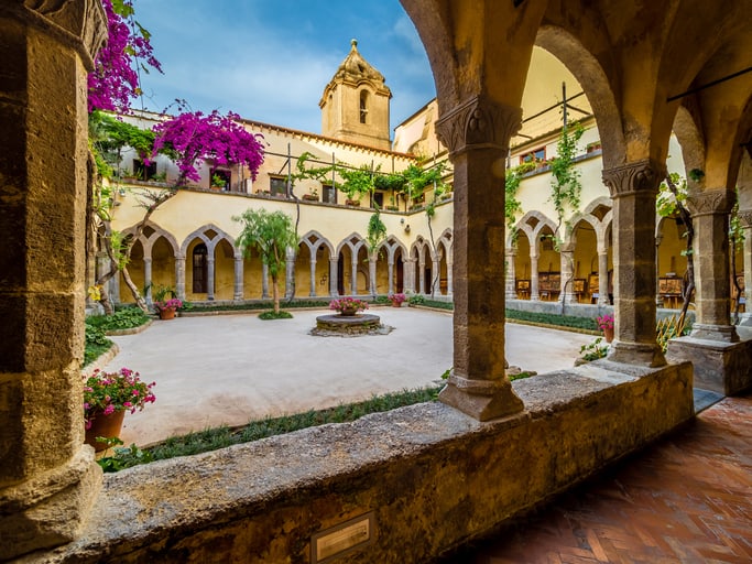 A serene courtyard, reminiscent of the cloister of San Francesco, boasts arches and climbing plants. It features a central well and is surrounded by vibrant flowers under a clear blue sky.