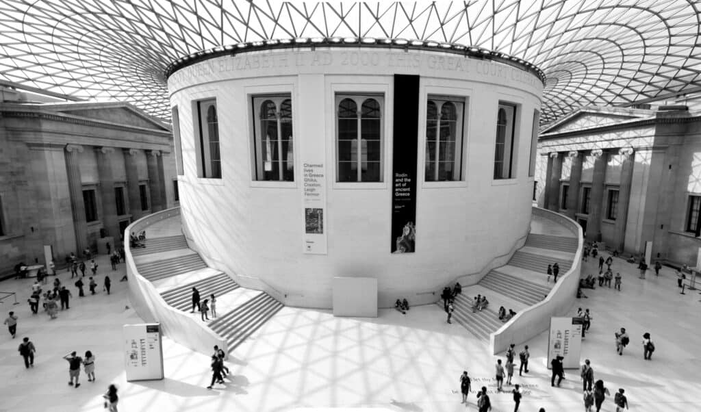 Interior view of a large, domed atrium at one of London's tourist attractions, with people walking on split staircases. It features a central round structure under a stunning geometric glass ceiling.