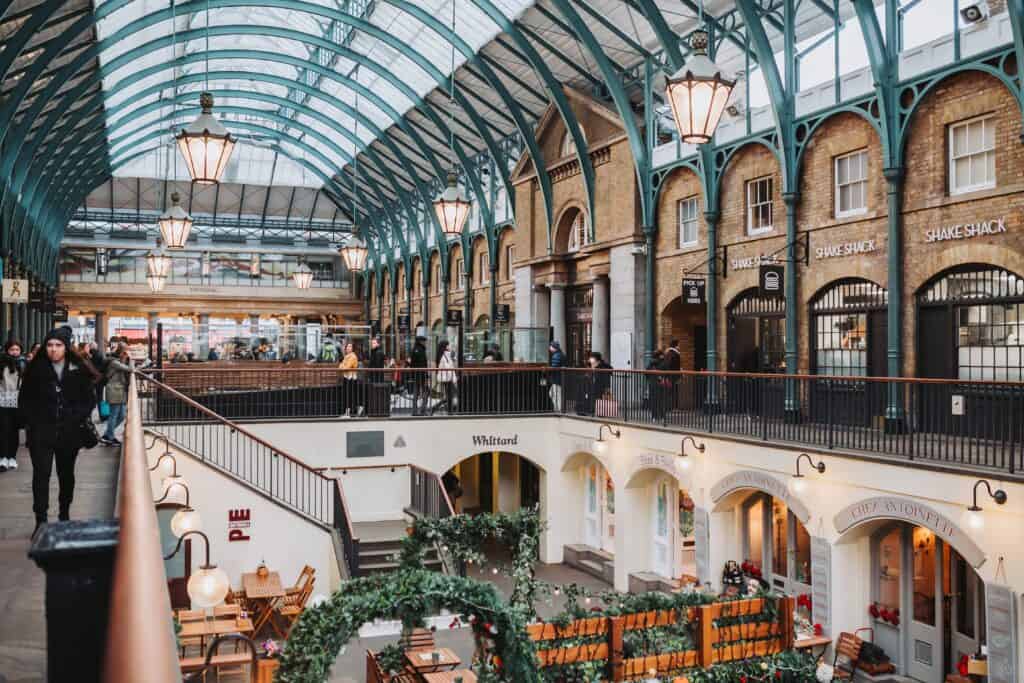 The bustling interior of this shopping area, one of the notable London tourist attractions, features an arched glass ceiling and multiple floors packed with diverse shops. Visitors wander through the stylish space, discovering unique finds as they embrace the vibrant city atmosphere.