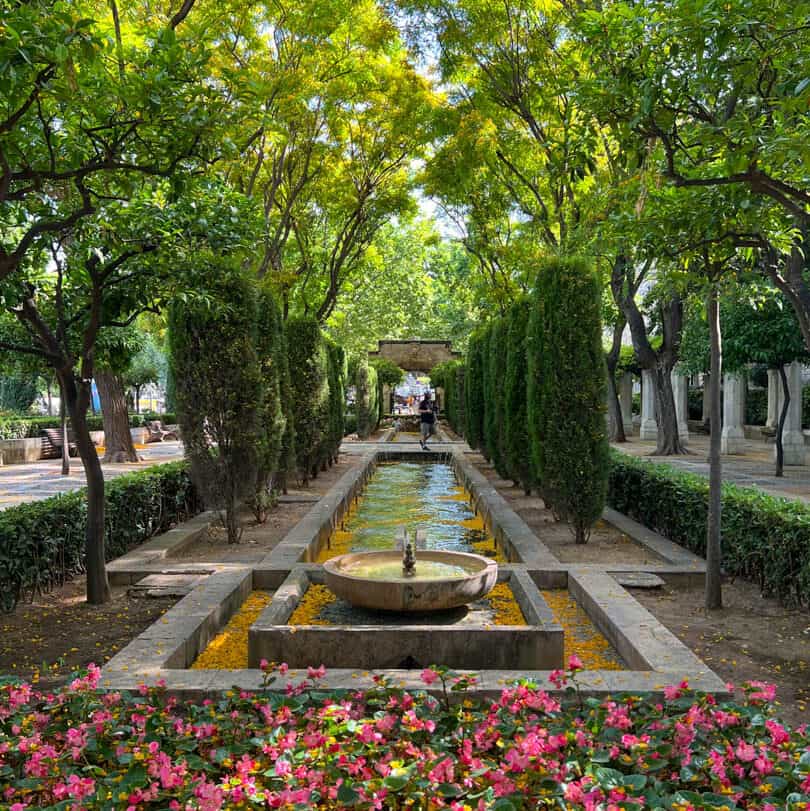 A stone fountain sits at the center of a rectangular water feature, flanked by trees, hedges, and pink flowers in a formal garden.