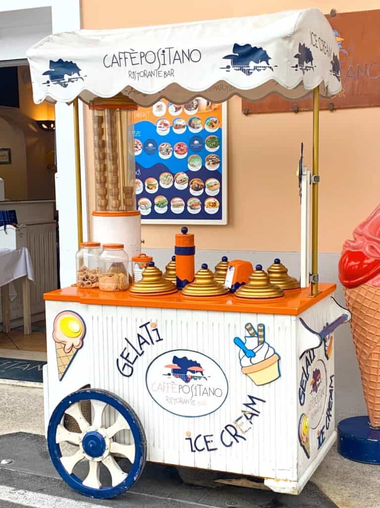 An ice cream cart labeled "Caffè Positano," adorned with cones, dispensers, and charming decorative elements reminiscent of the vibrant spirit of Positano, is parked on a bustling street.