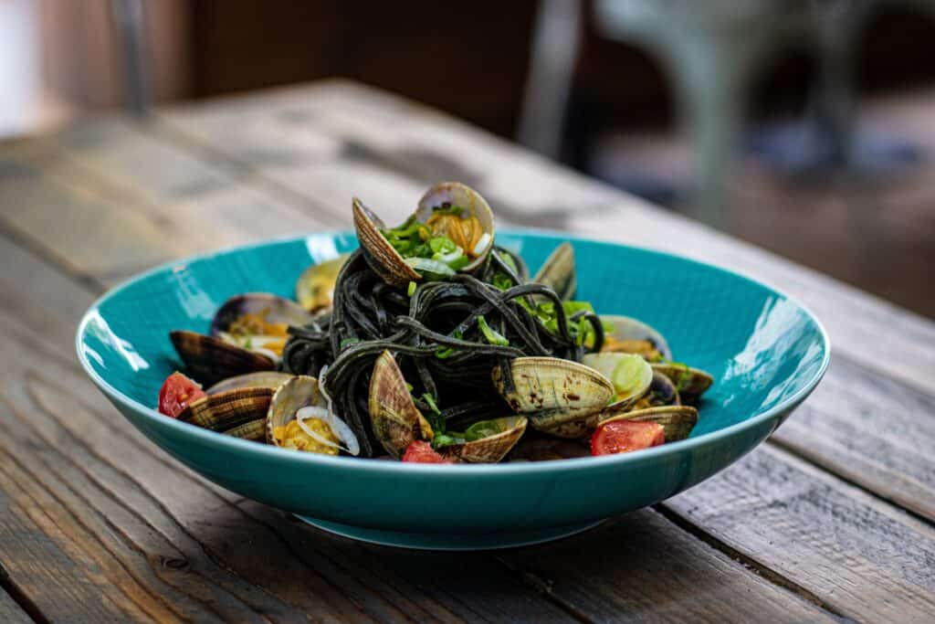 A blue bowl of squid ink pasta with clams, tomatoes, and greens sits on a wooden table, capturing the essence of coastal cuisine. Sampling this dish is one of the must-do things in Positano for a true taste of Mediterranean flavors.
