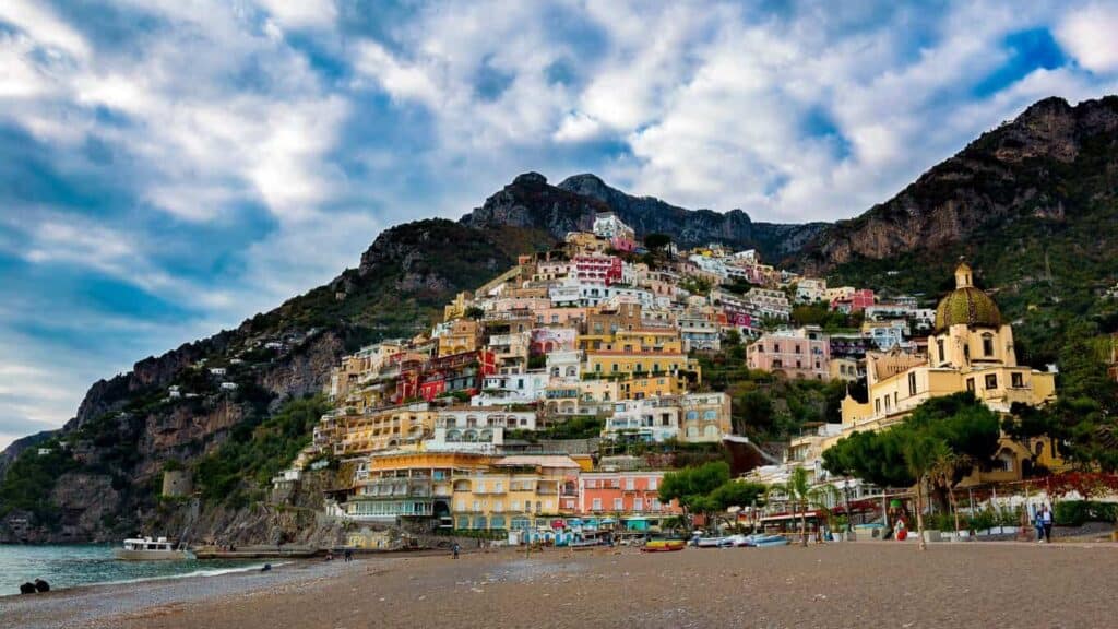 The enchanting hillside buildings of Positano, Italy, showcase a kaleidoscope of colors, with a beach and the shimmering sea in the foreground and a cloudy sky above.