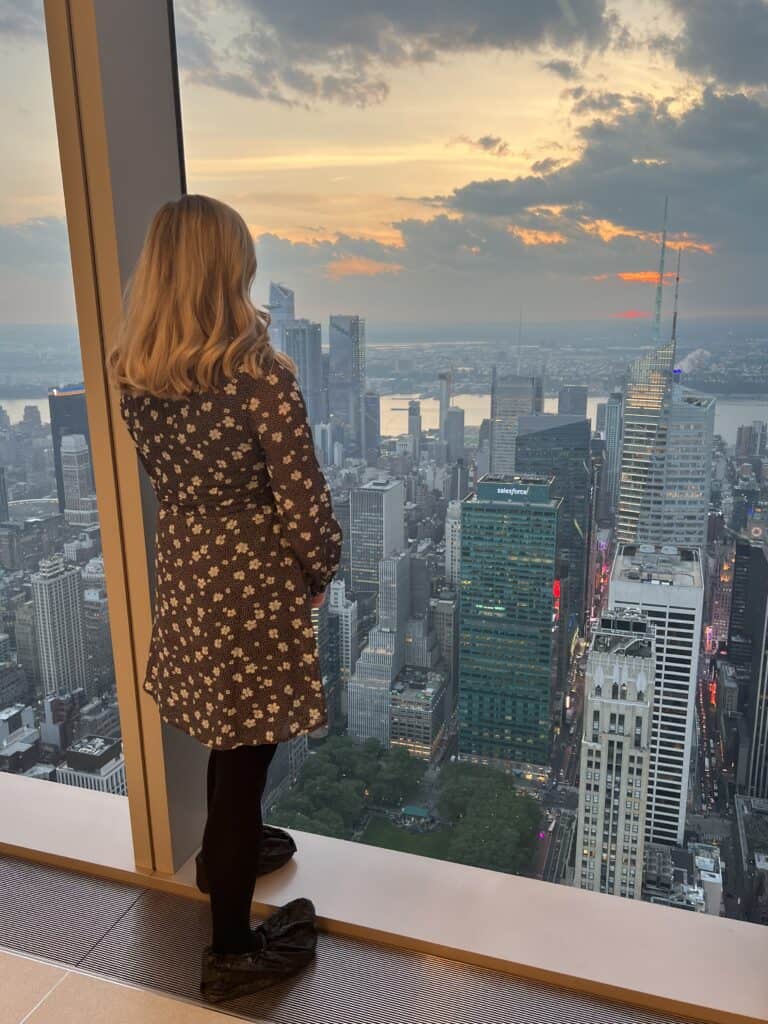 Woman with blonde hair stands at a floor-to-ceiling window looking out over New York City at sunset.