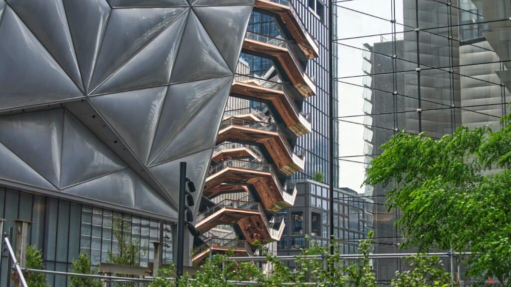 The image captures a modern architectural structure with geometric panels next to a tall, honeycomb-like building in Hudson Yards. Greenery flourishes in the foreground, offering a serene complement to one of the many things to explore in this vibrant area.