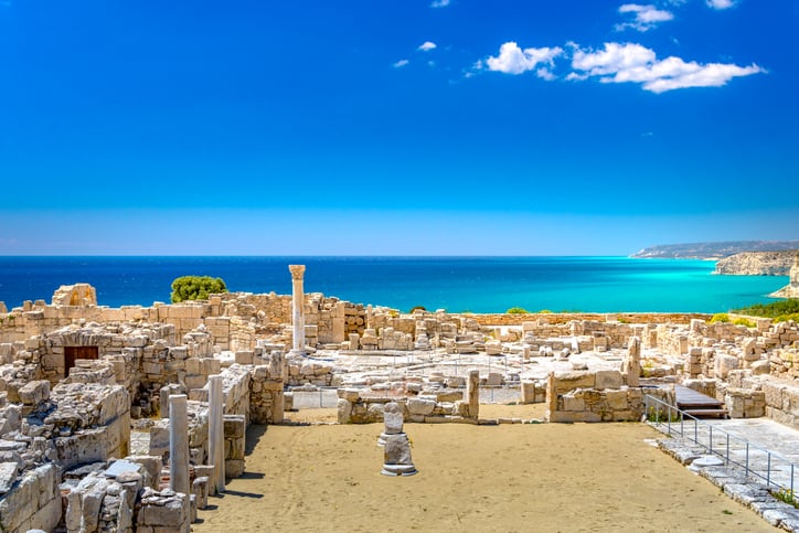Ancient stone ruins with columns stand near a sandy courtyard, overlooking a bright blue sea and clear sky.