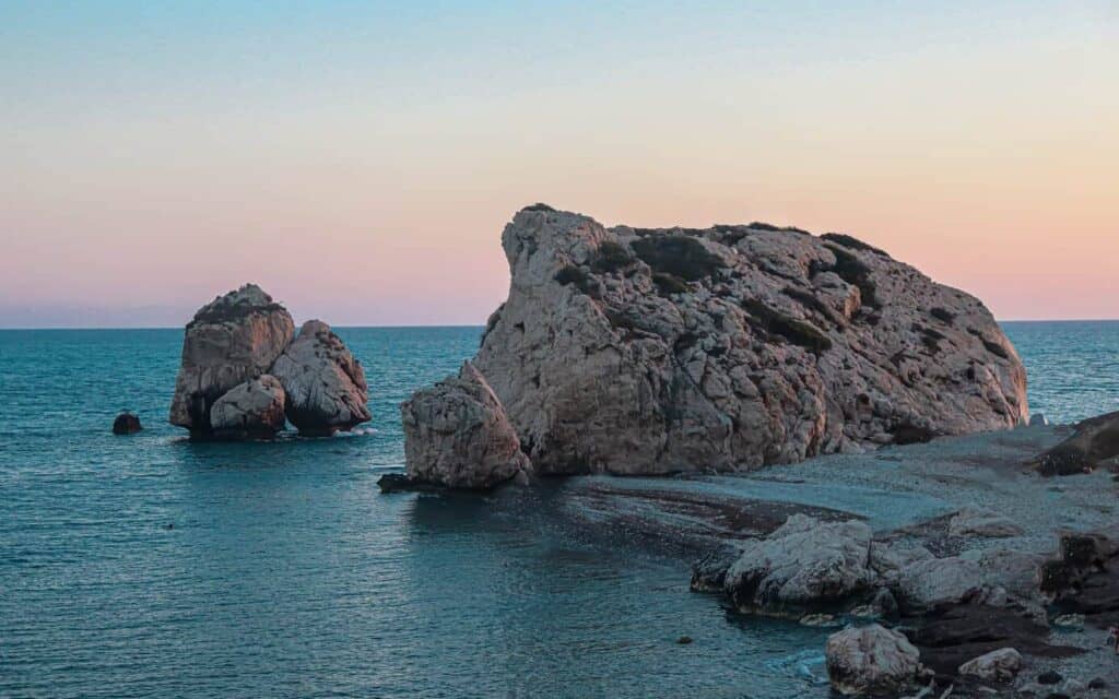 Large rock formations rise from the sea near the coastline at sunset, with calm blue water and a pastel sky in the background.