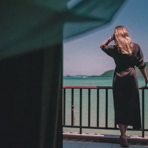 A woman in a dark dress stands on a balcony overlooking a calm sea and distant green hills under a clear sky.