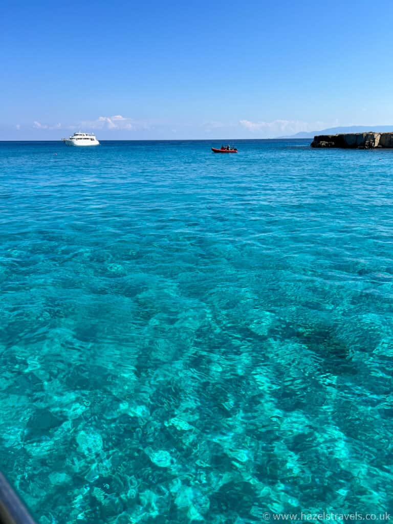 Explore the rocky coastline of Paphos under a clear blue sky, where the turquoise sea stretches out with a large white boat in the distance and a small red boat closer to shore.