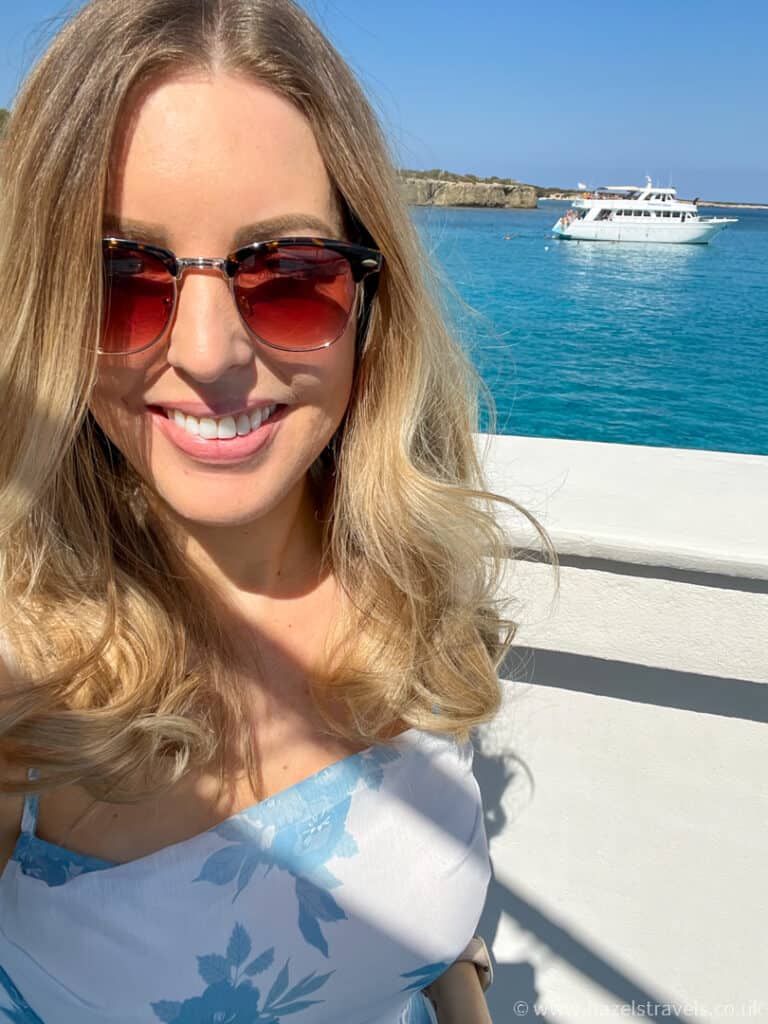Woman takes a selfie with blue turquoise water and a white boat in the background.