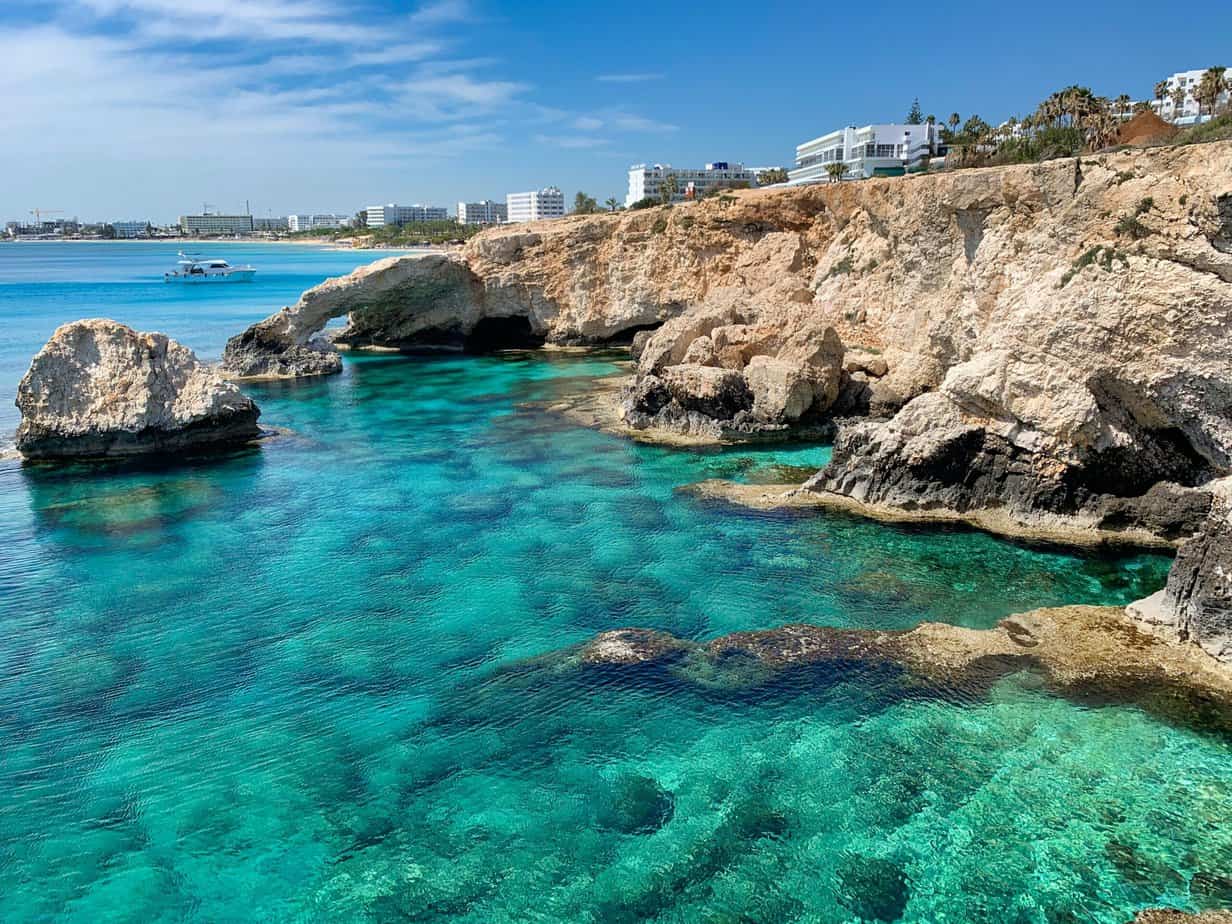Clear turquoise water and rocky cliffs under a blue sky, with buildings and a boat visible in the background—just some of the stunning sights to see in Cyprus.