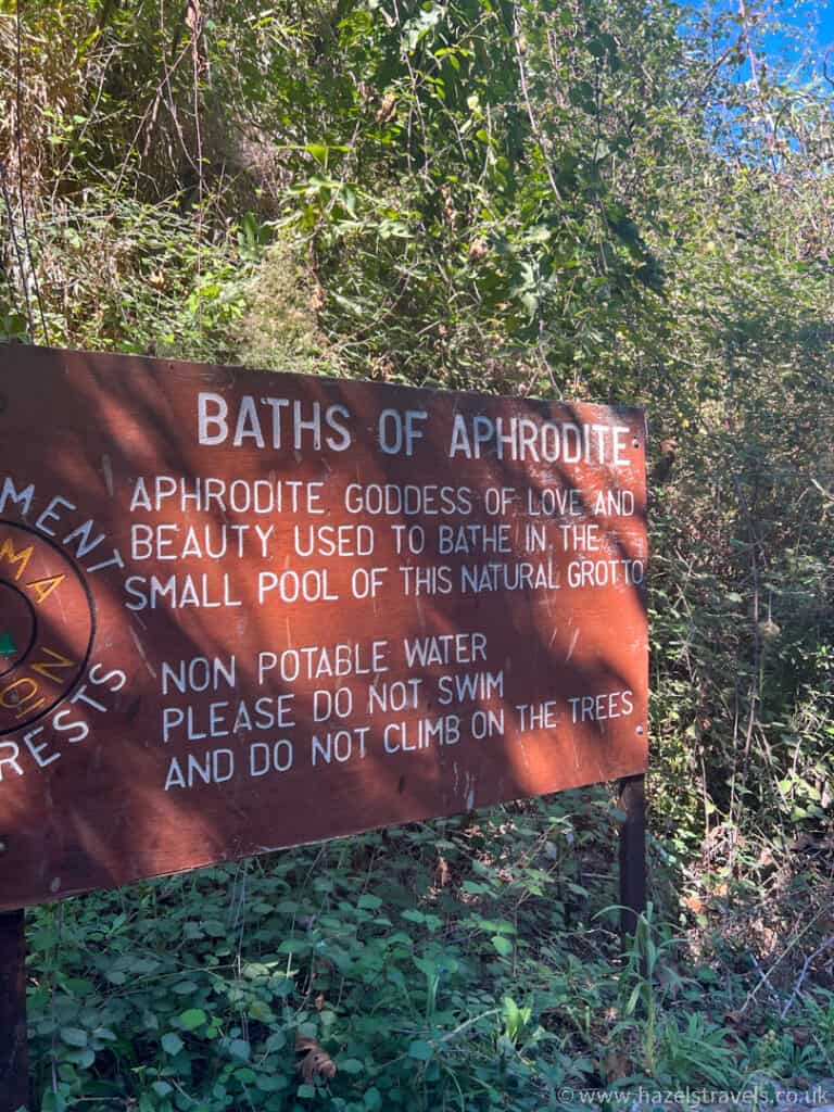 Brown sign in a wooded area reads “Baths of Aphrodite,” offering site details and warnings about non-potable water and tree climbing. Lush greenery surrounds this spot, making it a must-see among things to do in Paphos.