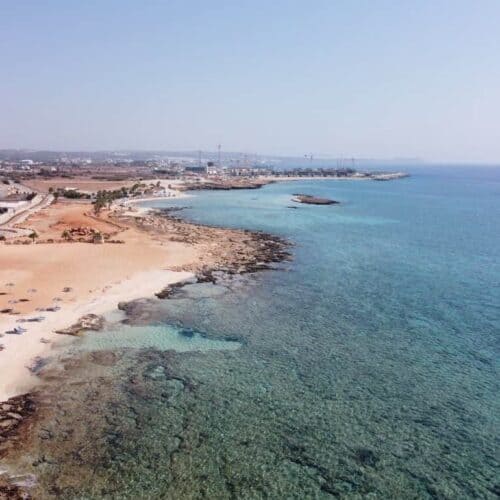 Aerial view of a sandy beach with umbrellas, clear blue water, and nearby buildings along the coastline.
