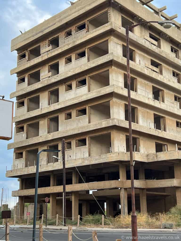 A multi-story, unfinished concrete building with exposed beams and no windows stands near the Famagusta border crossing, surrounded by overgrown weeds and a deserted street.