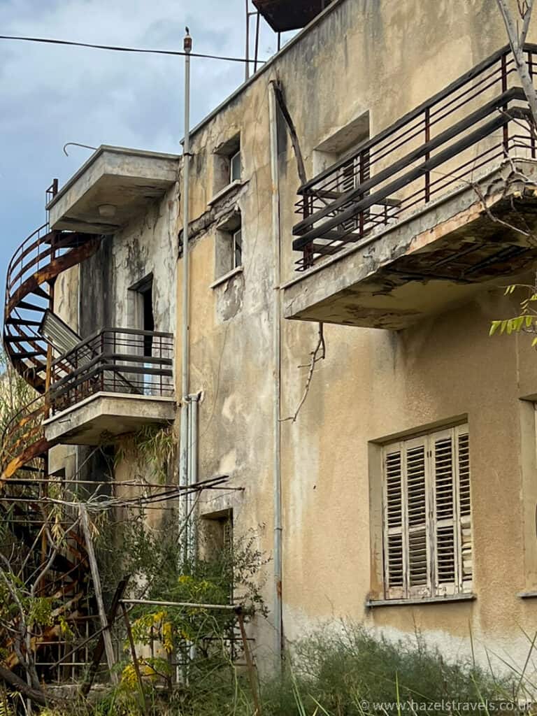 A weathered, abandoned building with peeling paint, rusted spiral staircase, broken railings, and overgrown vegetation