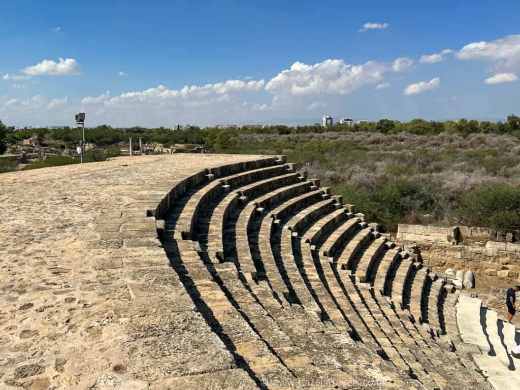 An ancient stone amphitheater with tiered seating is pictured outdoors, surrounded by greenery under a blue sky with scattered clouds