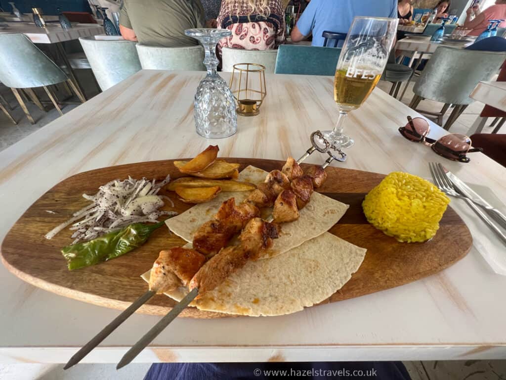 A wooden platter with grilled meat skewers on flatbread, yellow rice, roasted potatoes, salad, a green pepper, and a glass of beer awaits you at a restaurant near the Famagusta border crossing.
